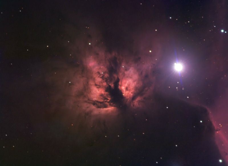 Orange-pink nebula split by dark dust lanes next to a very bright star at right, with dark nebulosity below