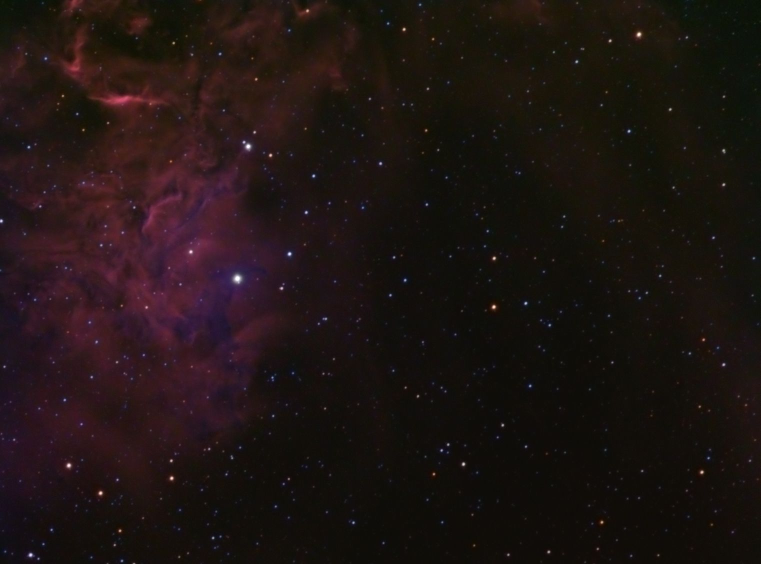 Wispy red and purple emission nebulosity along the left side of a dark star field