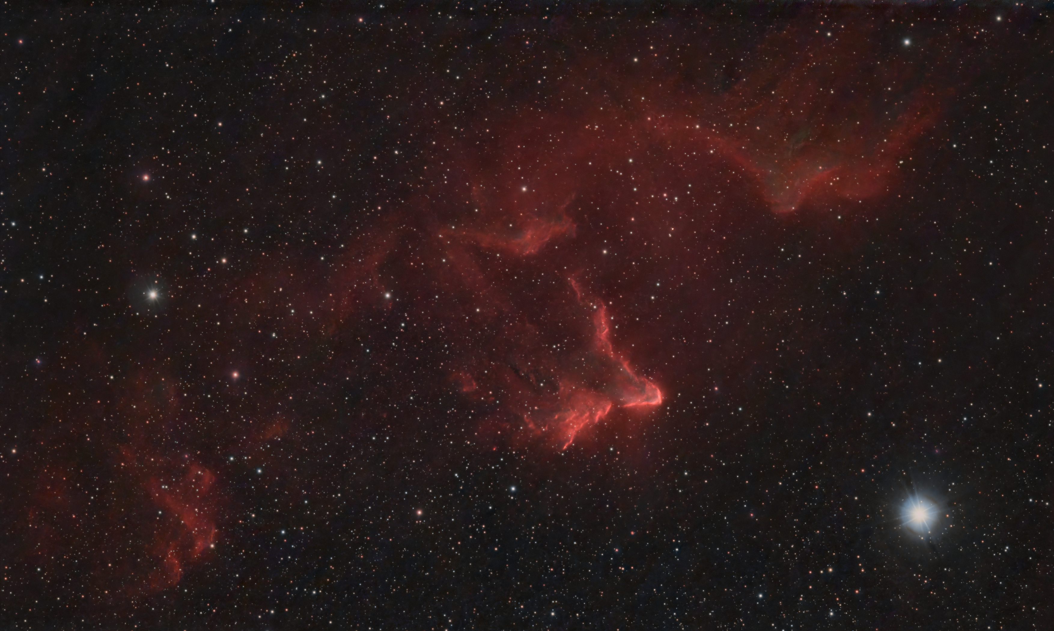 Red glowing emission nebula with a bright rim, shaped like a ghost, in a star field with a bright blue-white star at lower right