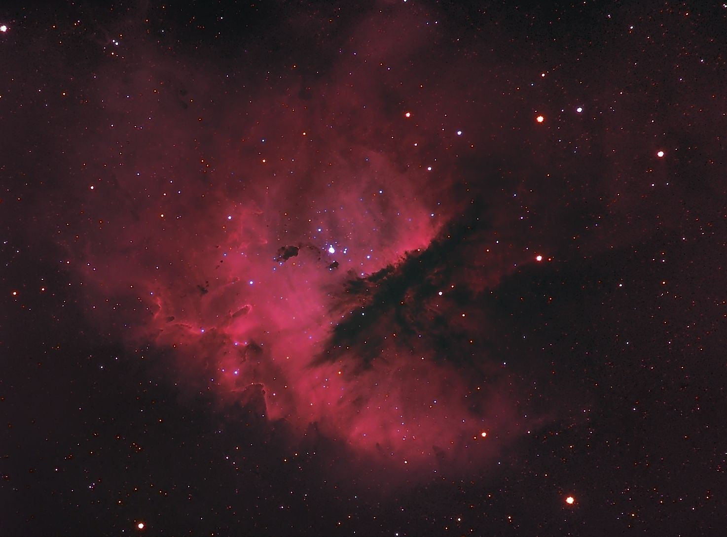 Pink-red emission nebula with a dark dust lane cutting in from the lower right and a cluster of young stars at its centre