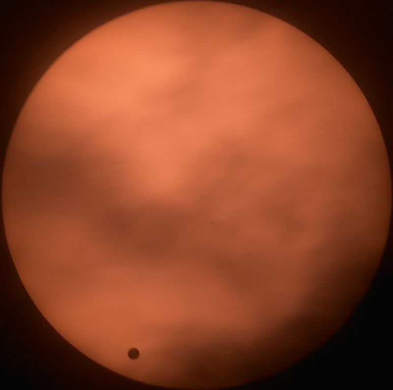 Orange disc of the Sun seen through thin cloud, with the small black disc of Venus near the lower left edge