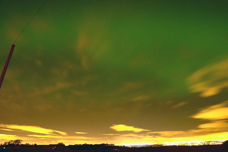 Green aurora filling the sky above orange-lit clouds and distant town lights