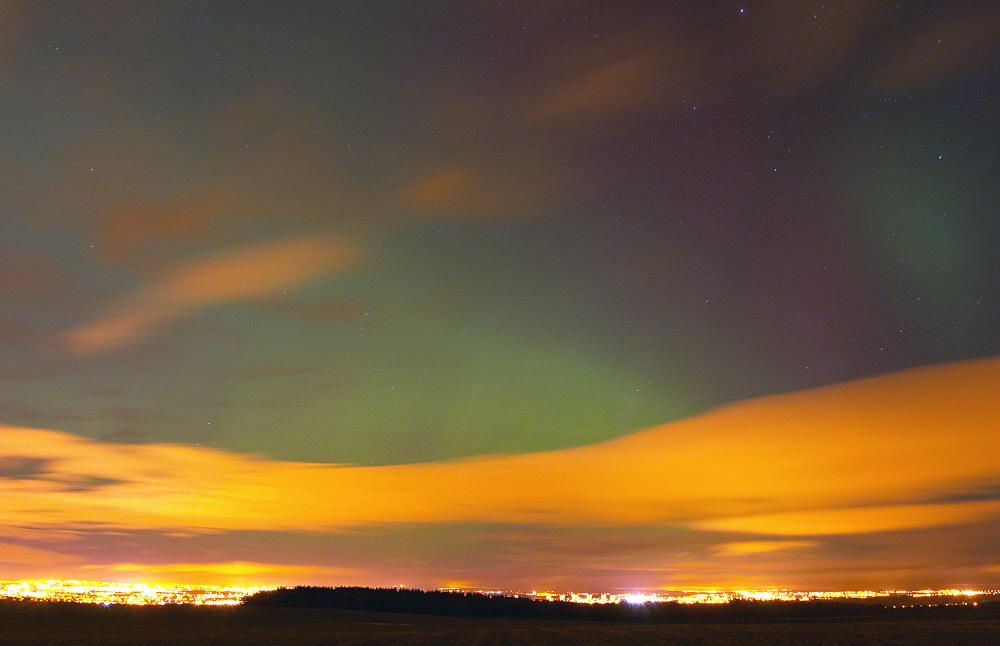 Faint green and red aurora behind bands of orange-lit cloud over a low horizon