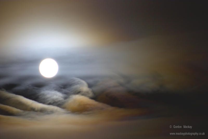 Bright Moon surrounded by a soft coloured glow above rolling clouds